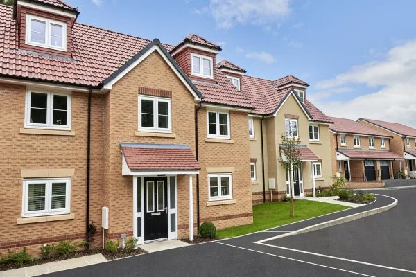 A row of modern, two-story brick houses by Elan Homes features red-tiled roofs and dormer windows. Each house boasts a black front door, white-framed windows, and a small front garden. The street is clean and bordered by neatly trimmed grass. Spencer Cobby