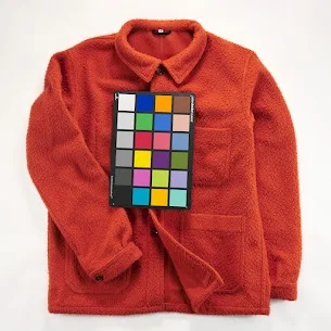 A vibrant orange fleece jacket lays flat on a white surface, with a color calibration chart showcasing various swatches, enhancing the colored images. The jacket boasts a collar and front pockets, offering both style and functionality. Spencer Cobby