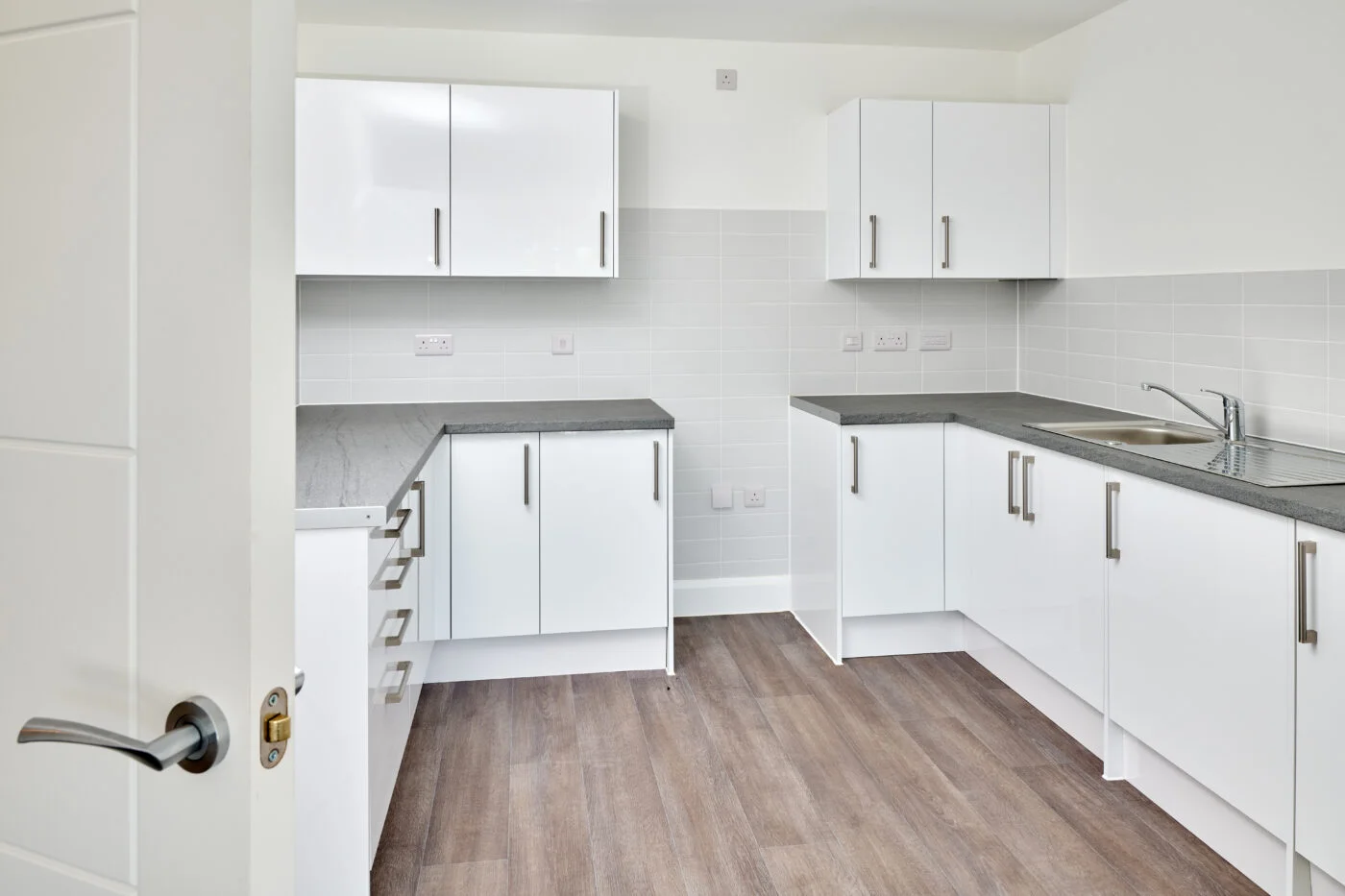 Modern kitchen with white cabinets, light gray countertops, and a tiled backsplash. The room has wood-style flooring, a stainless steel sink, and a partially open door on the left. Spencer Cobby