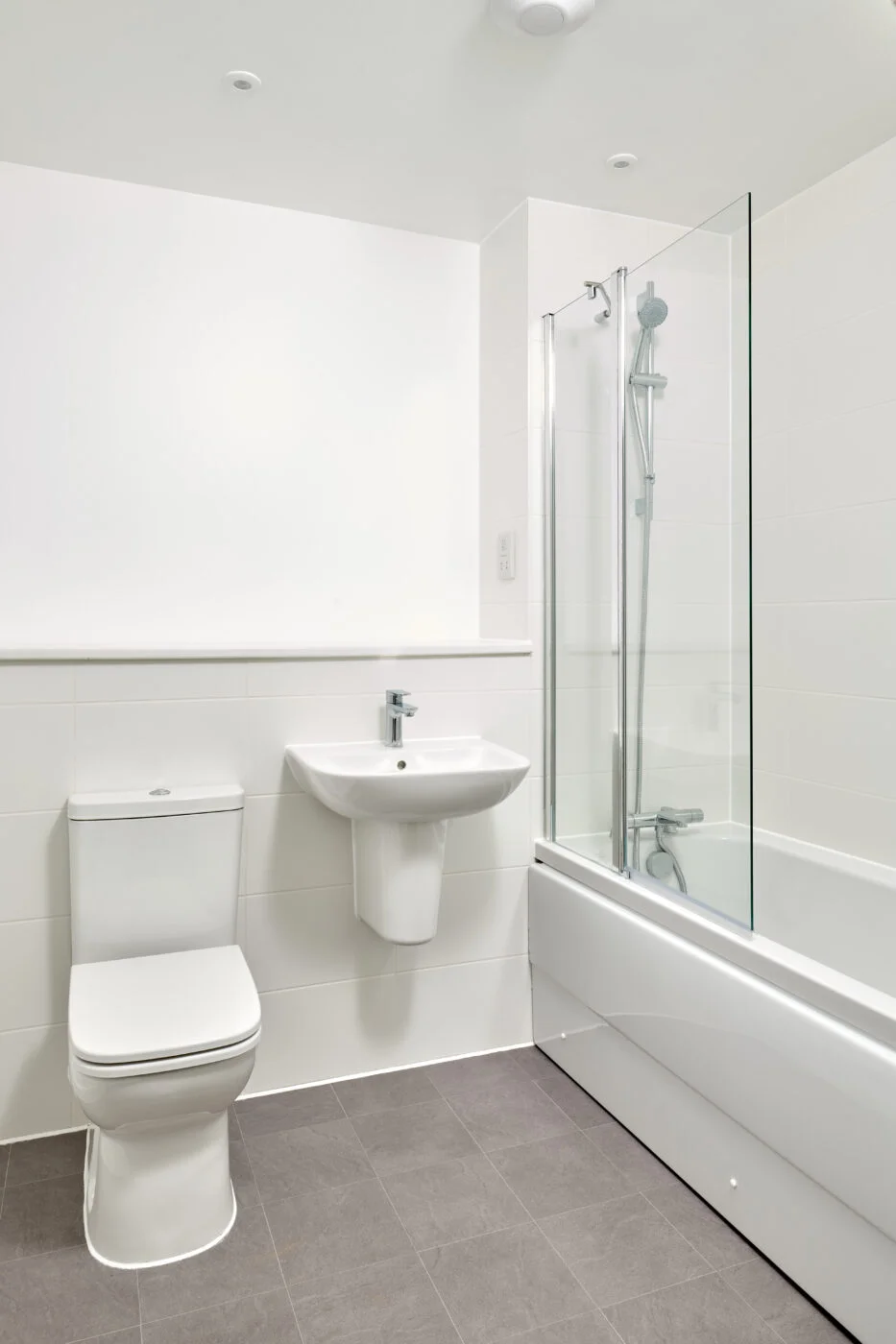 A modern, white bathroom featuring a wall-mounted sink, toilet, and a bathtub with a glass shower screen. The walls are tiled, and the floor has gray tiles, giving the space a clean, minimalistic look. Spencer Cobby