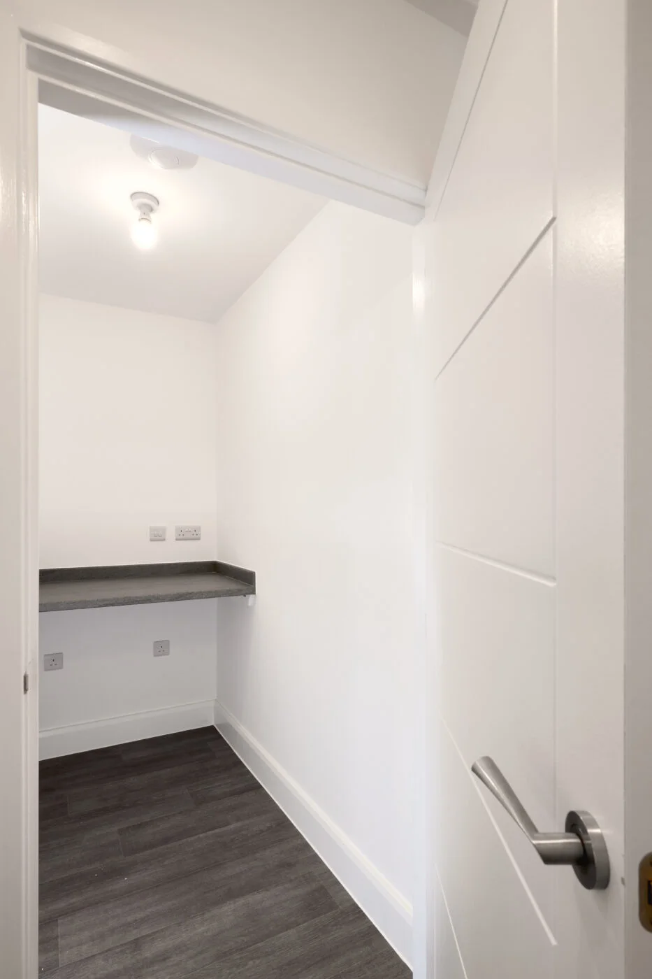 A small, empty utility room with plain white walls, a single ceiling light, and a dark countertop built into the wall. The room has dark wood flooring and multiple electrical outlets near the countertop. The door is white with a silver handle. Spencer Cobby