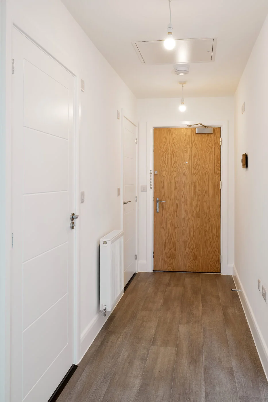 A minimalist hallway with a wooden floor and white walls. A wooden door with a metal handle is at the end, flanked by white panel doors and a radiator on the left. Overhead lighting softly illuminates the space. Spencer Cobby