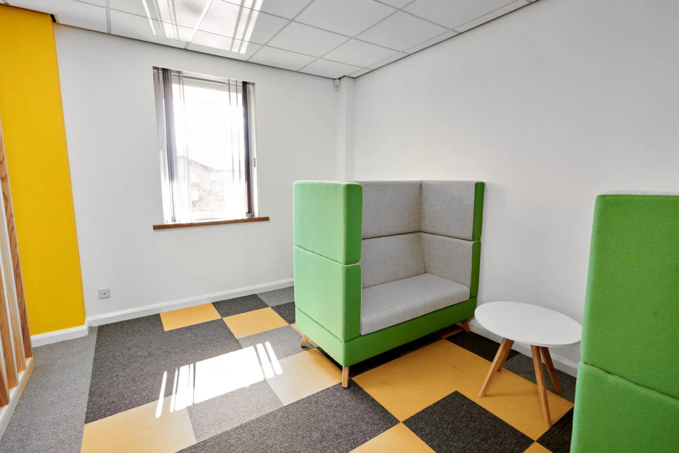 A modern office space with a gray and yellow checkered carpet. There’s a green upholstered booth seating with a small white round table. A window with vertical blinds lets in natural light, and the walls are white with a yellow accent. Spencer Cobby