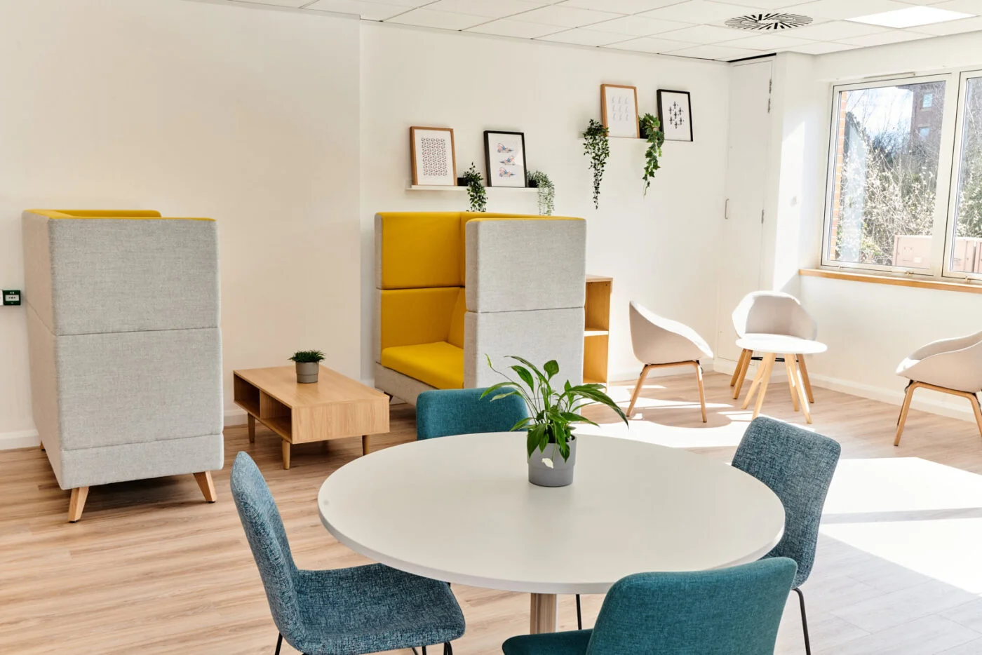 A modern office lounge with natural light. It features a round white table with blue chairs, two gray and yellow high-back seats, small wooden tables, framed art on the wall, and a window with sunlight streaming in. Spencer Cobby