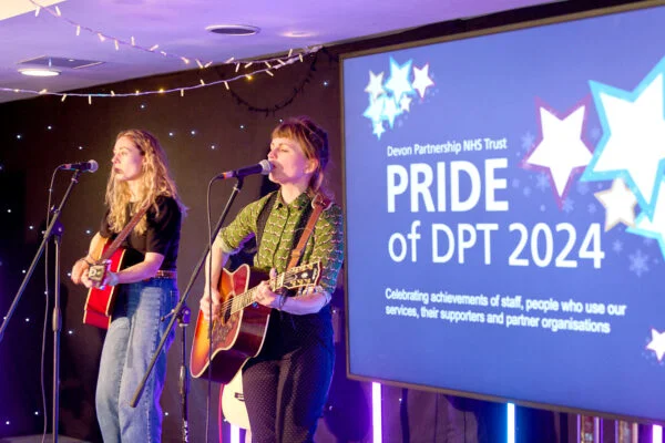 Two musicians perform on stage with guitars at an event. A screen displays "Devon Partnership NHS Trust PRIDE of DPT 2024" and a message celebrating achievements and community support. Star decorations and string lights enhance the ambiance. Spencer Cobby