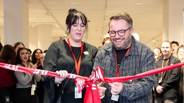 A man and woman smiling as they cut a red ribbon with scissors, surrounded by a cheering crowd in a brightly lit room. Both are wearing lanyards and appear excited, suggesting an opening event. Spencer Cobby