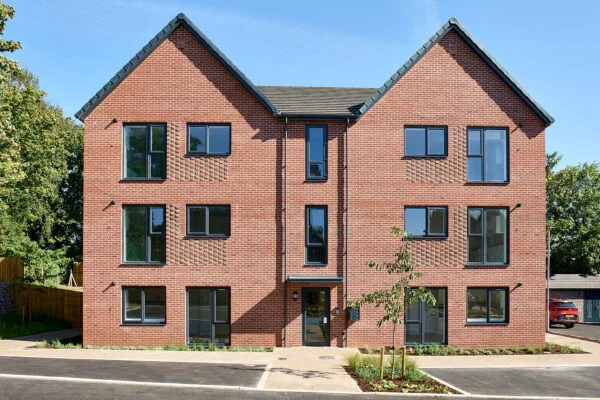 A modern three-story brick apartment building with dark-framed windows under a blue sky. A small tree is planted in front, and the surrounding area features a paved walkway and greenery. Spencer Cobby