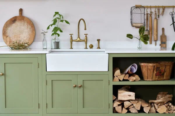 A kitchen with green cabinets and a farmhouse sink. There's a brass faucet, wooden countertop decor, and a plant in a vase. Firewood and a basket are stored in open shelves. Hanging utensils are visible above. Spencer Cobby