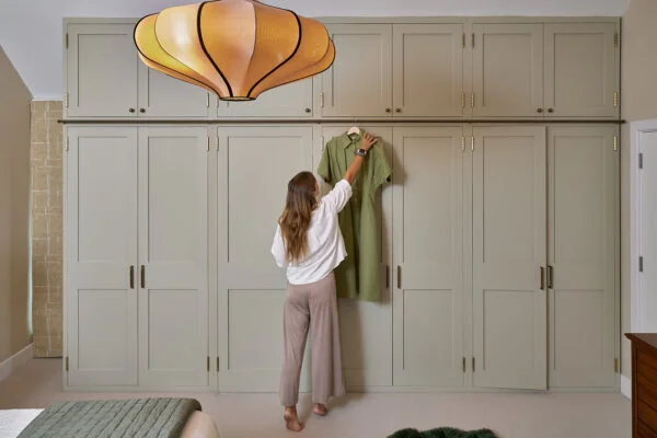 A person reaches to hang a green dress on a sage green wardrobe cabinet in a bedroom. The room features a large pendant light, neutral tones, and a neatly made bed with a green blanket. Spencer Cobby