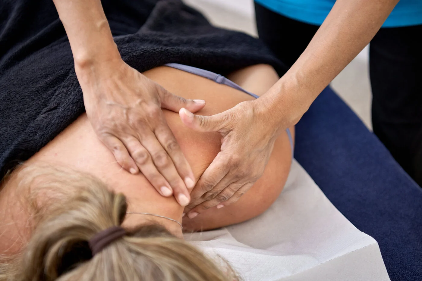 A person receiving a shoulder and back massage on a massage table. The person is lying face down, covered with a dark towel. The massage therapist uses both hands to apply pressure to the shoulder area. Spencer Cobby