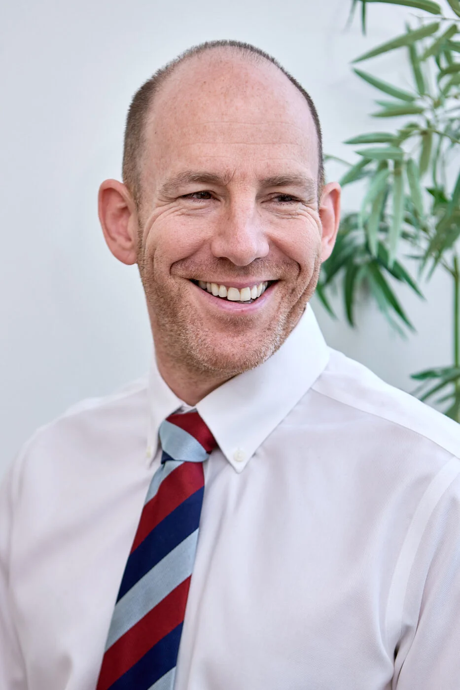 A smiling man with a shaved head and a striped tie looks to the side. He is wearing a white shirt, and there's a leafy plant in the background. Spencer Cobby