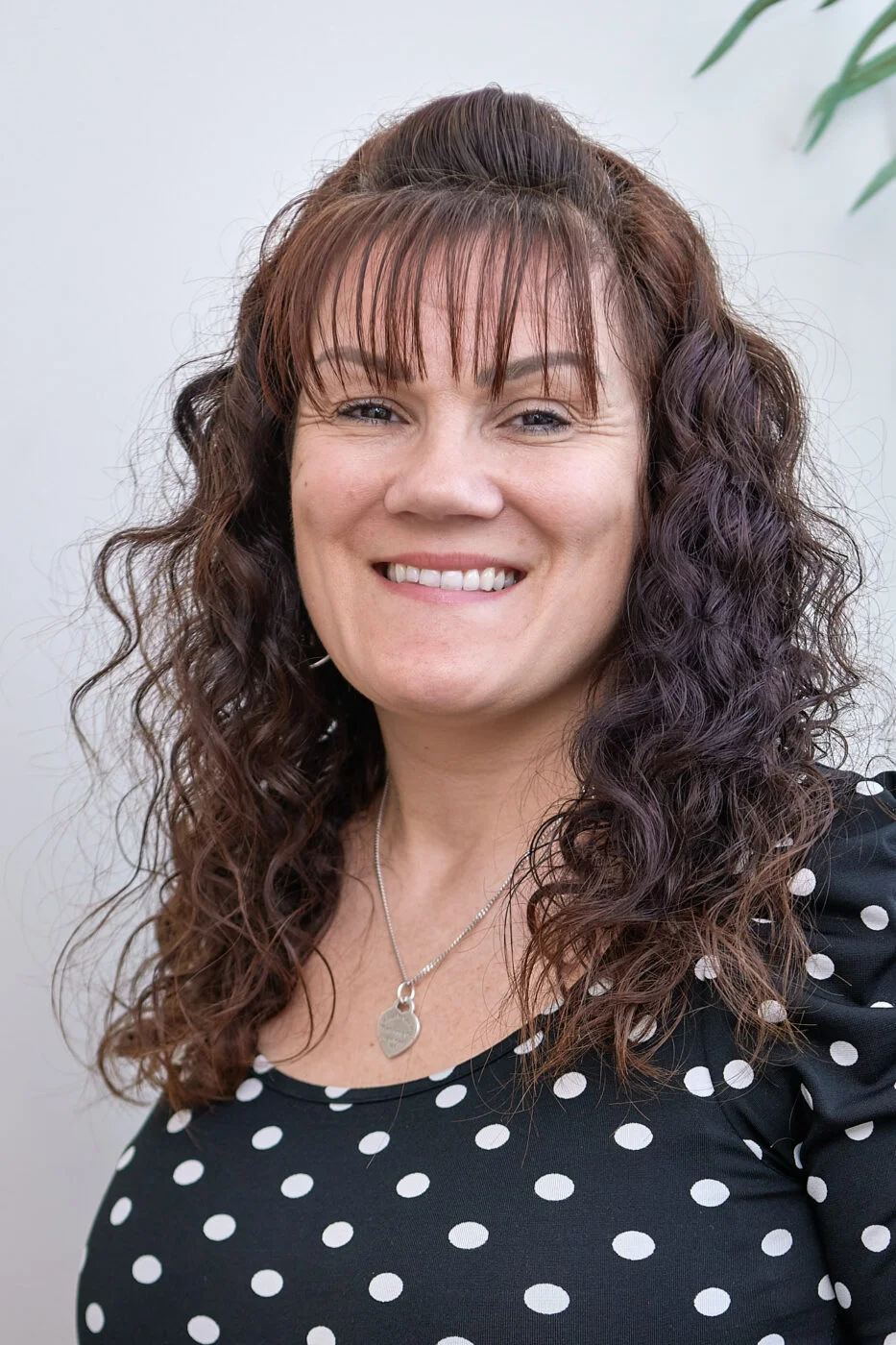 A person with curly brown hair smiles standing against a plain background. They are wearing a black shirt with white polka dots and a necklace. Spencer Cobby