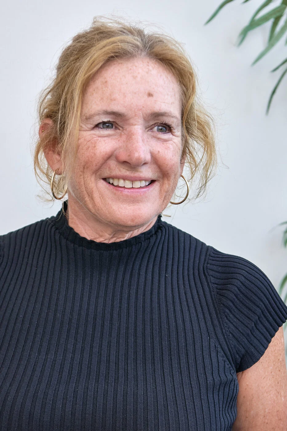 A woman with light brown hair and freckles is smiling. She is wearing a black ribbed top and hoop earrings. The background is a soft white with a partial view of a green plant. Spencer Cobby