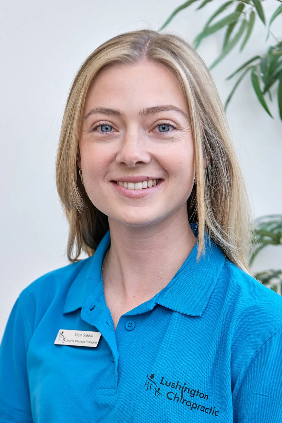 A woman with long blonde hair is smiling. She is wearing a blue polo shirt with a "Lushington Chiropractic" logo and a name tag. There is a green plant in the background. Spencer Cobby