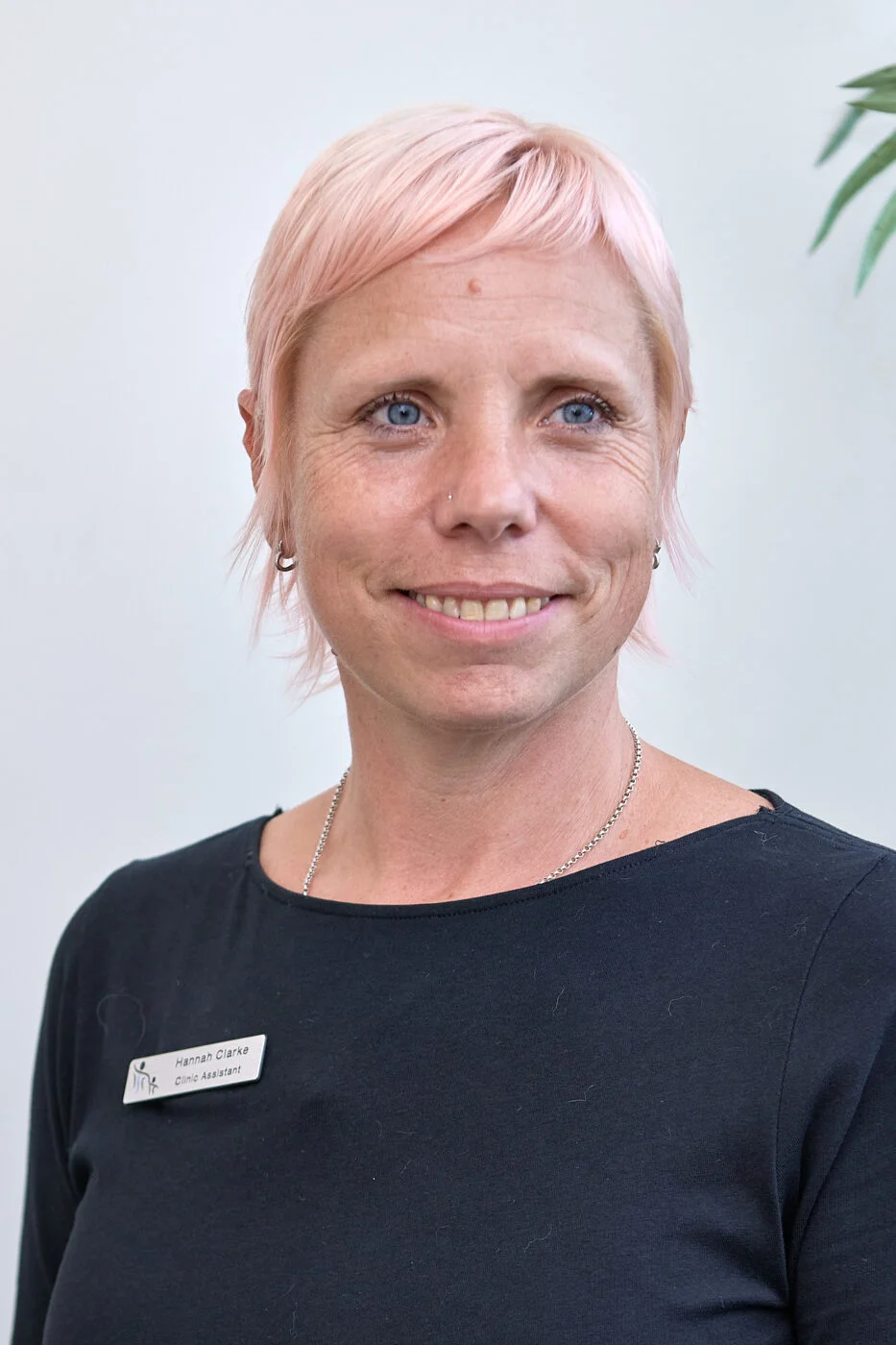 A person with short pink hair is wearing a black top and a name tag. They are standing in front of a light-colored background and looking slightly to the side with a neutral expression. Spencer Cobby