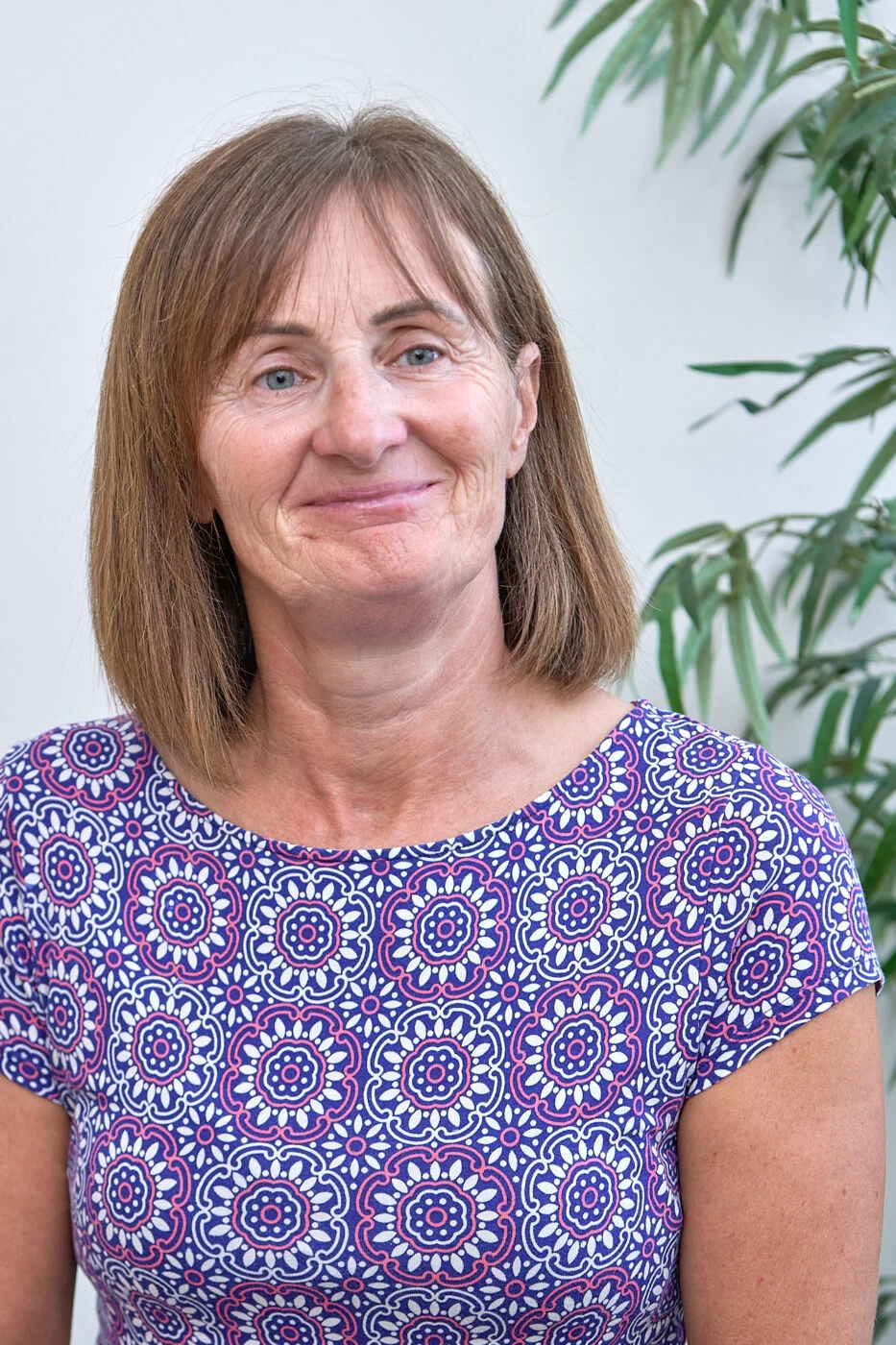 A woman with shoulder-length brown hair smiles while wearing a patterned purple top. She stands in front of a white background with green plant leaves partially visible beside her. Spencer Cobby