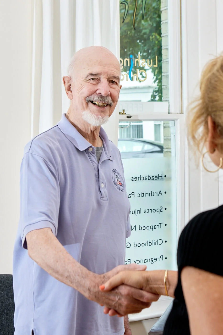 An elderly man with a white beard wearing a light purple polo shirt smiles and shakes hands with a person whose back is turned to the camera. They stand in a bright room with a window displaying text. Spencer Cobby