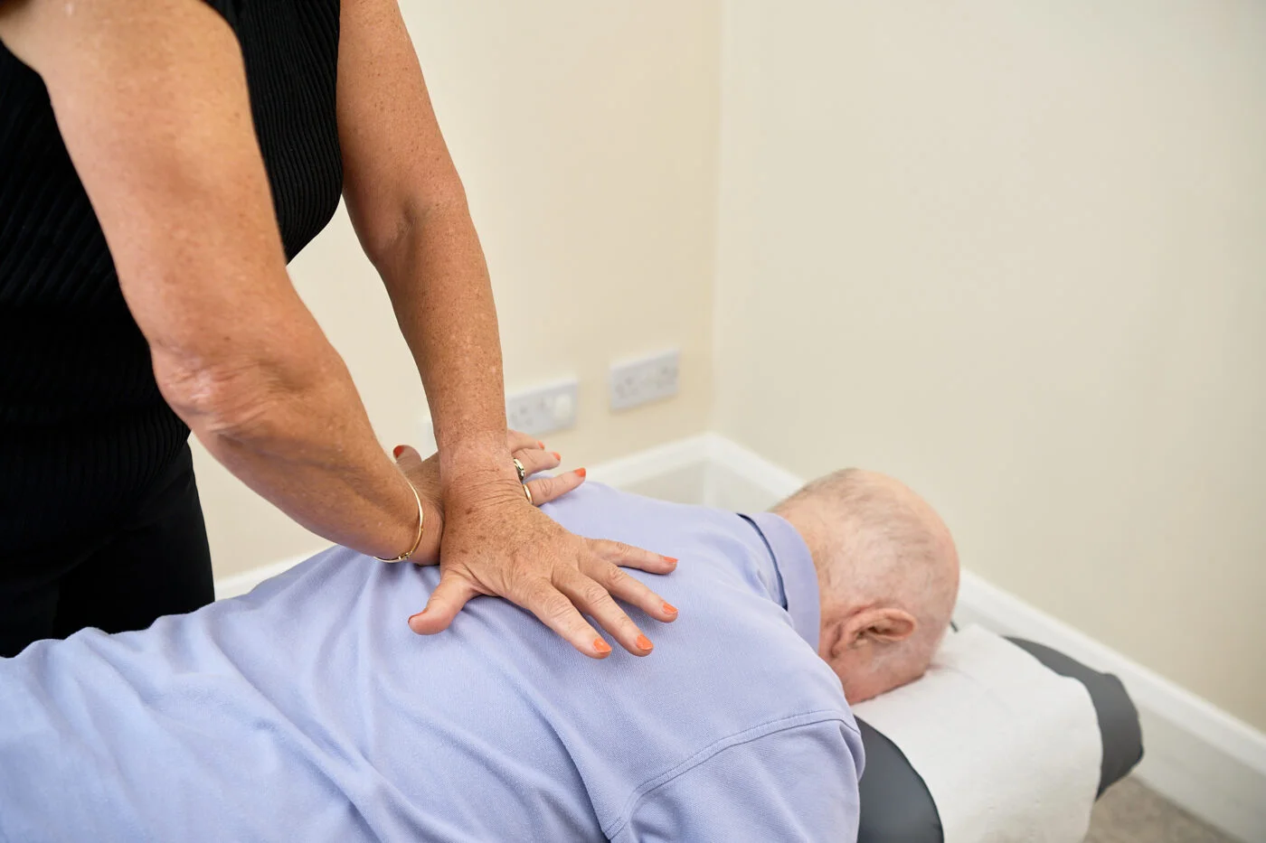 A person receiving a back massage while lying face down on a massage table. A therapist stands to the side, applying pressure with both hands on the person's back. The room has light walls and electrical outlets visible on the wall. Spencer Cobby