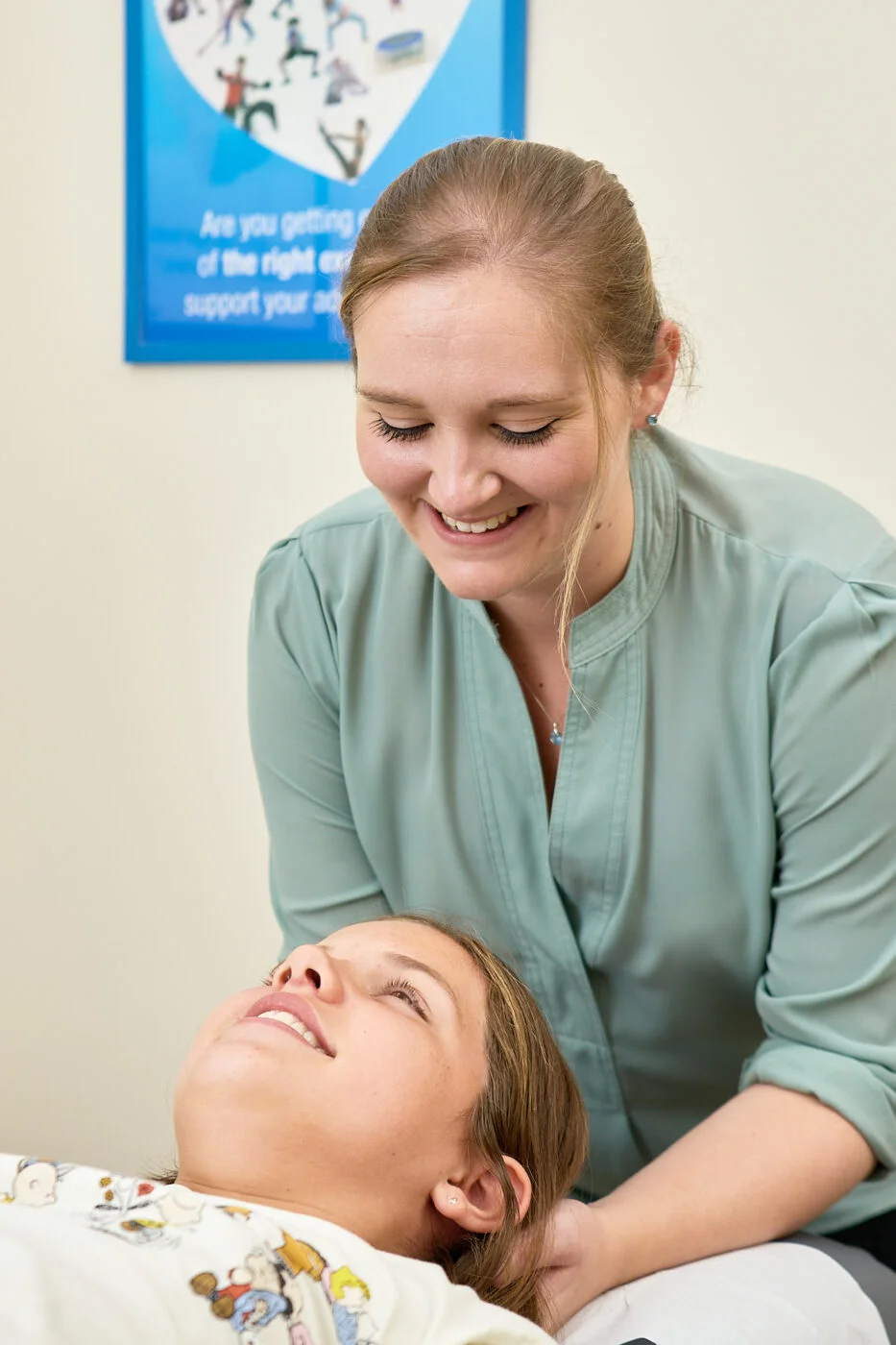 A woman in a green blouse smiles down at a girl lying on a bed during a therapy session. The room has a blue poster on the wall in the background. Spencer Cobby