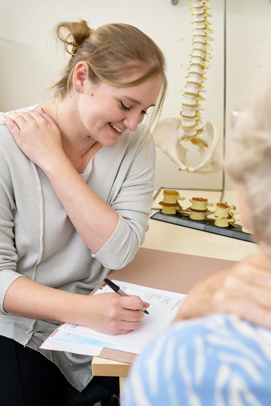 A woman smiles while writing on a clipboard, engaging in a consultation. A skeletal model of a spine is visible in the background. The setting suggests a medical or therapeutic environment. Spencer Cobby