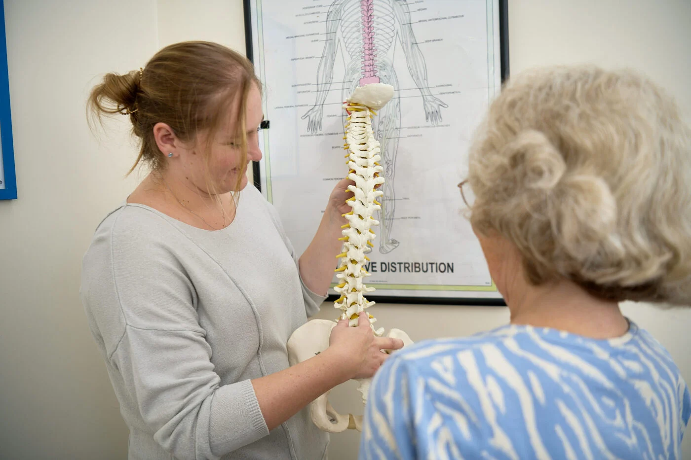 A healthcare professional shows a spine model to a patient in an office setting. A poster with spinal information is on the wall in the background. Spencer Cobby