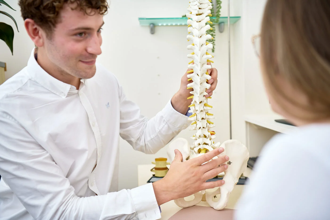 A person in a white shirt shows a spine model to another individual in an office setting. The focus is on the anatomical model, highlighting the vertebrae and pelvis. Shelves and a plant are visible in the background. Spencer Cobby