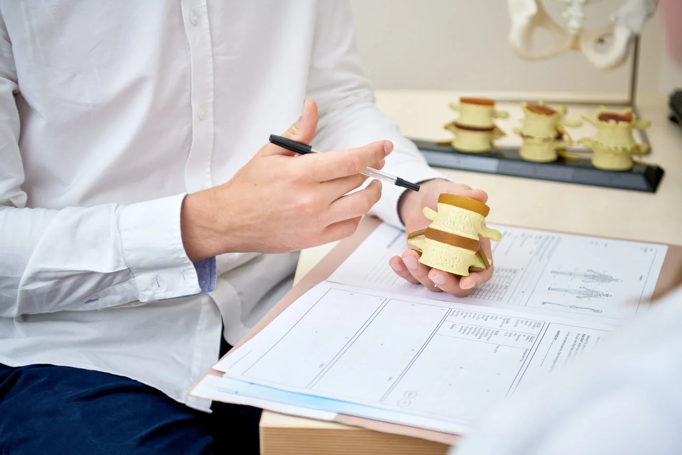 A person in a white shirt holds a spine model and points to it with a pen, discussing anatomical details. Open documents and diagrams are spread on the table, with more spine models visible in the background. Spencer Cobby