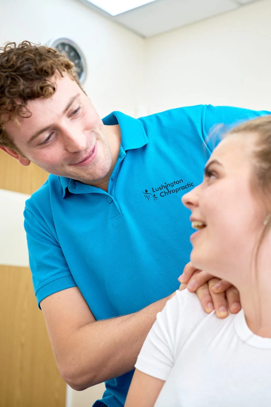 A chiropractor in a blue polo shirt is performing an adjustment on a smiling woman sitting in a clinic room. The chiropractor is gently pressing her shoulder. A wall clock is visible in the background. Spencer Cobby