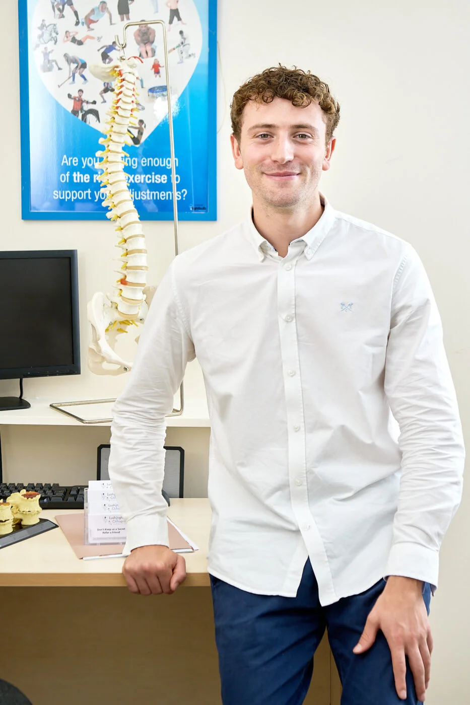 A person in a white shirt stands smiling in an office next to a spine model. A computer and anatomical models are on the desk, with a spine poster on the wall in the background. Spencer Cobby