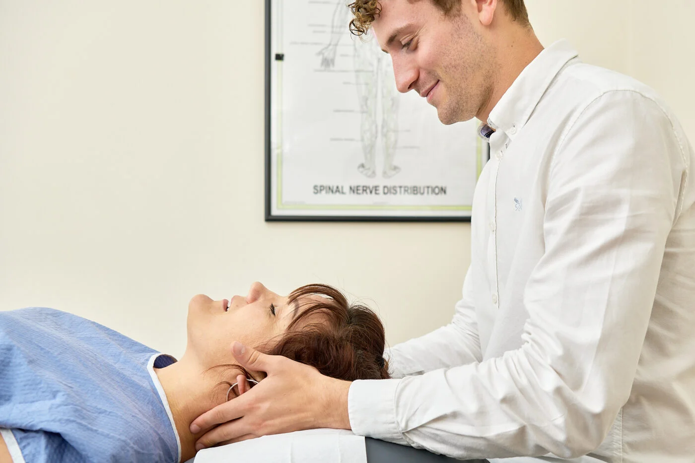 A chiropractor, wearing a white shirt, gently adjusts the neck of a woman lying on an examination table. A poster of spinal nerve distribution is on the wall in the background. Spencer Cobby