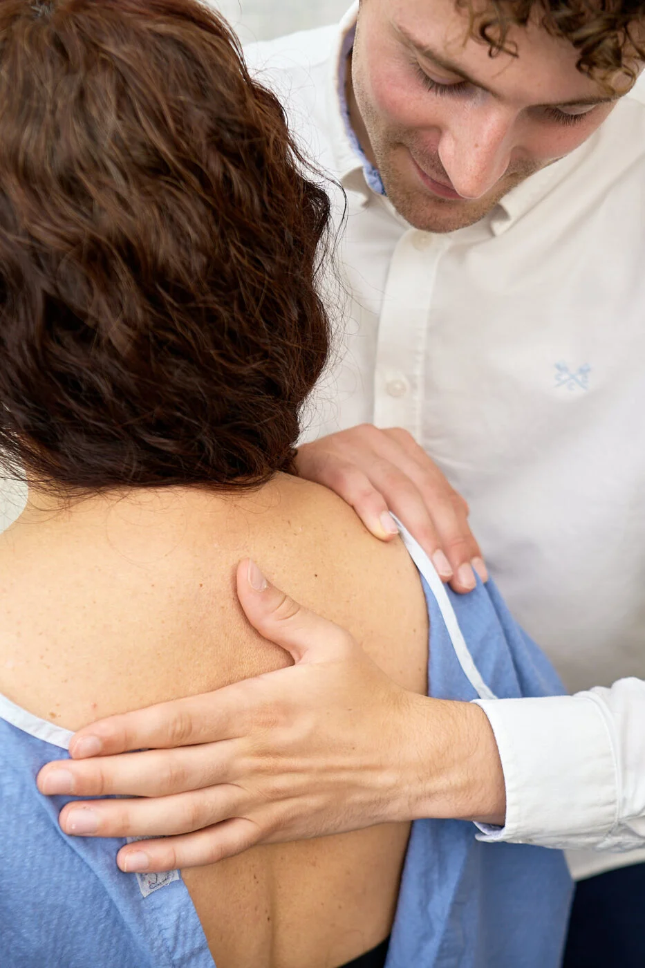 A person wearing a white shirt places their hand on the back of another person wearing a blue medical gown. The scene appears to be in a clinical or medical setting. Spencer Cobby