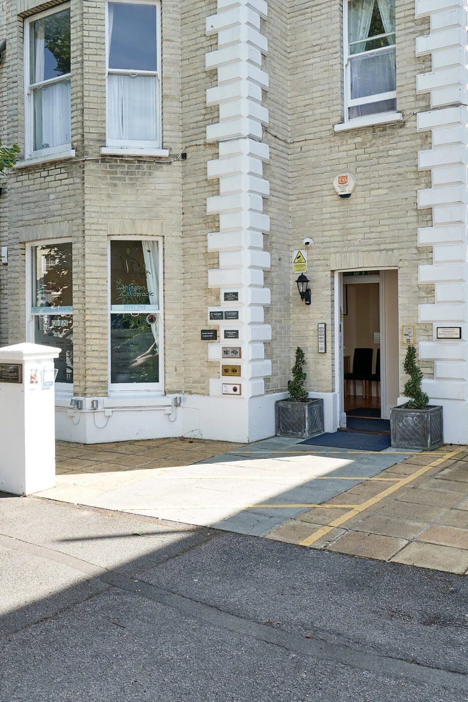 Entrance to a brick building with signs and potted plants on either side of the door. The wall is light-colored with several windows, and a yellow line is marked on the pavement nearby. Spencer Cobby