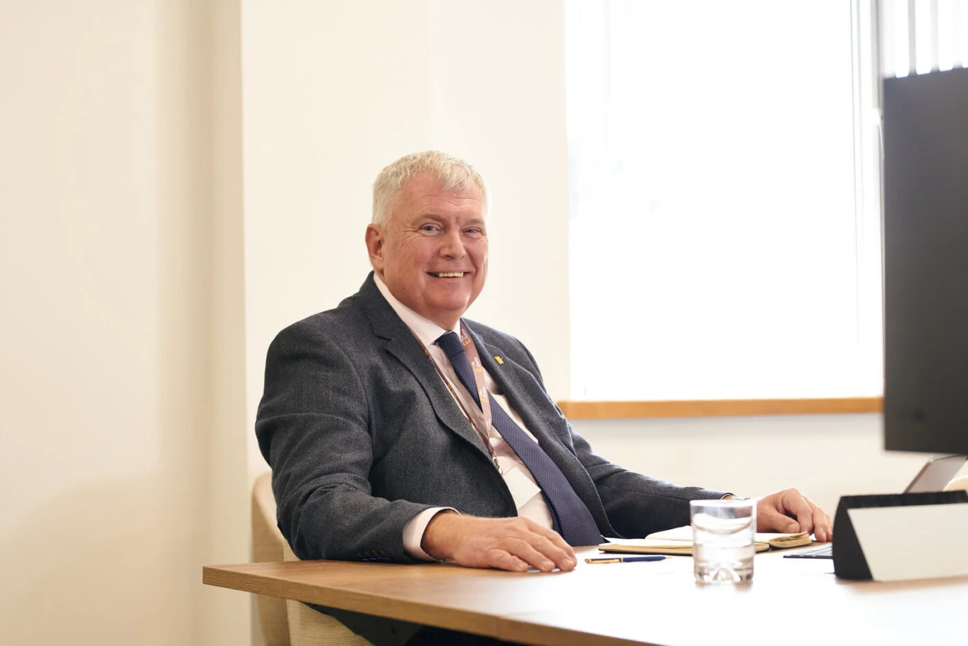 An older man with white hair sits at a desk, smiling while wearing a dark suit and tie. He faces the camera with a glass of water and a notebook on the table. Soft light filters through a window behind him, giving the room a bright ambiance. Spencer Cobby