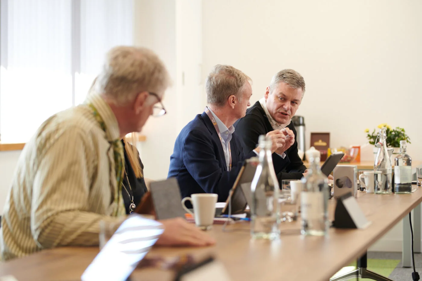 Three men are seated at a conference table engaged in discussion. They have notebooks, water bottles, and a coffee mug on the table. The background shows a white wall and window with a vase of yellow flowers. Spencer Cobby