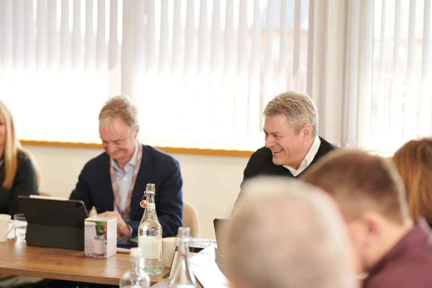 A group of people sits around a table during a meeting in a well-lit room. Two men in focus are smiling and using electronic devices. The table has bottles, papers, and a box. Blinds cover the windows in the background. Spencer Cobby