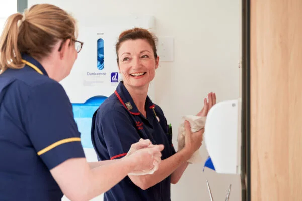 Two healthcare workers in navy uniforms are smiling and talking while washing their hands in a medical facility. One holds paper towels, and a dispenser is mounted on the wall nearby. Spencer Cobby