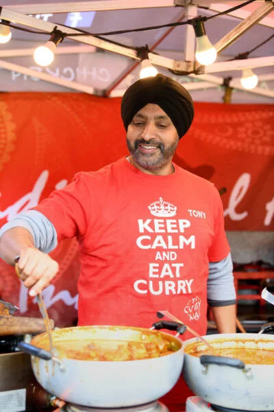 A man wearing a turban and a red shirt that says "KEEP CALM AND EAT CURRY" is serving curry from large pots at a food stall. He is smiling and holding a ladle. The background has festive lighting and a red banner. Spencer Cobby