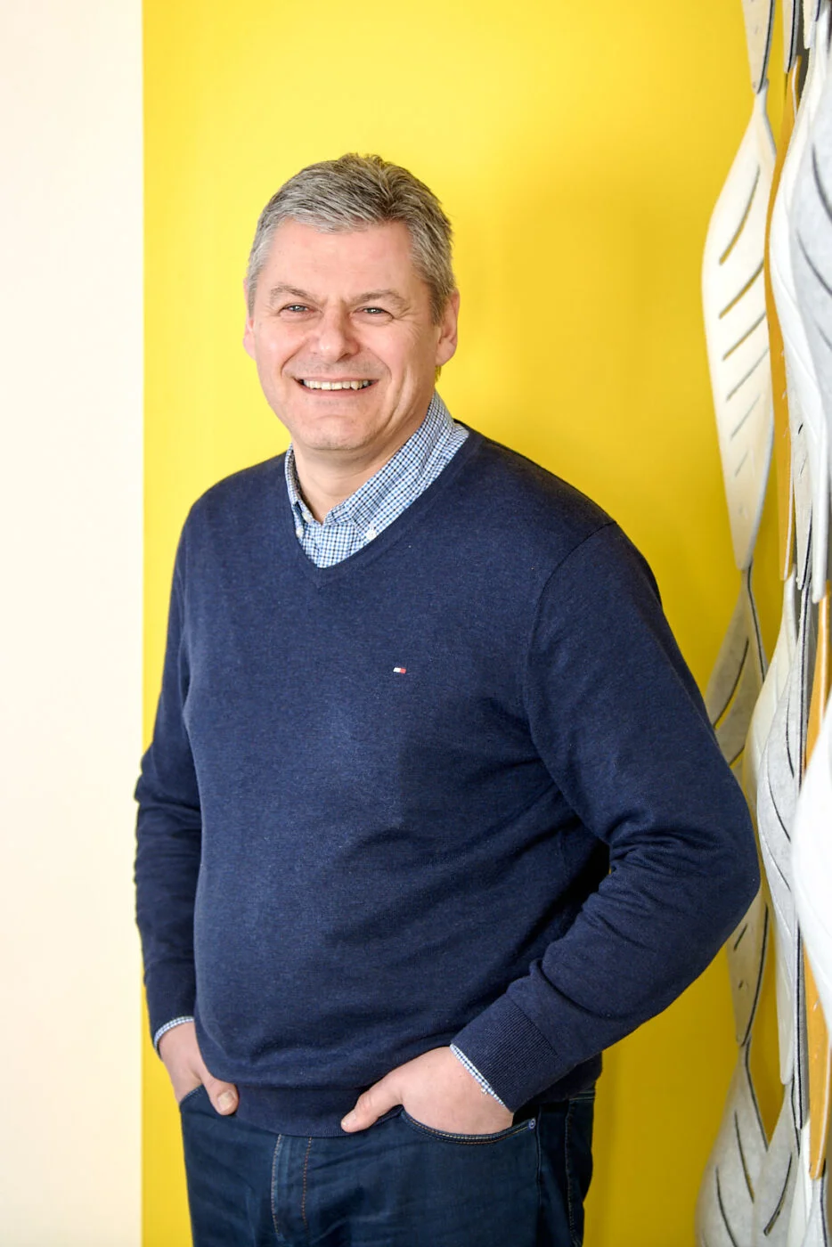 Smiling man wearing a blue sweater and jeans stands against a bright yellow wall with decorative elements on the side. Spencer Cobby