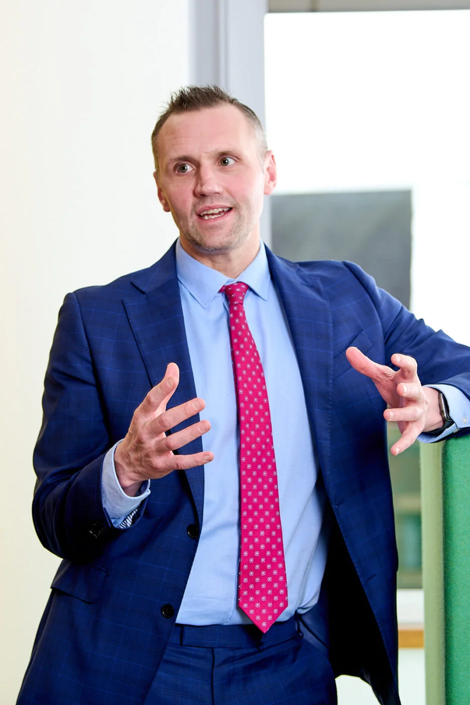 A man in a blue suit and pink polka dot tie is speaking and gesturing with his hands. He is standing indoors, leaning against a green surface. Spencer Cobby