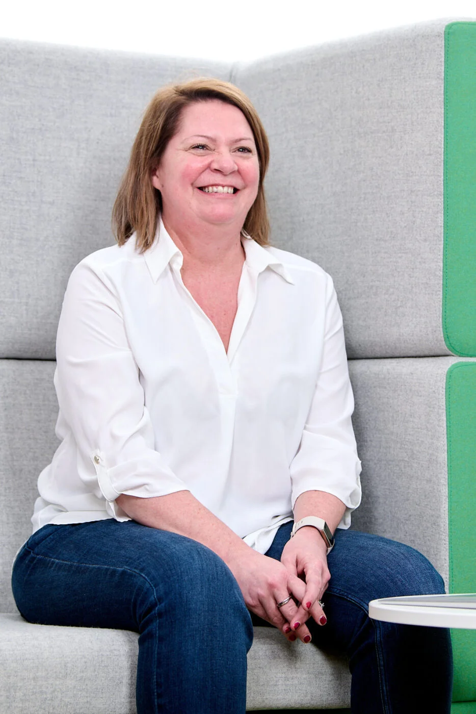 A woman with shoulder-length brown hair is sitting on a light gray sofa. She is wearing a white blouse and blue jeans, smiling and looking ahead. The background features soft gray and green upholstery. Spencer Cobby