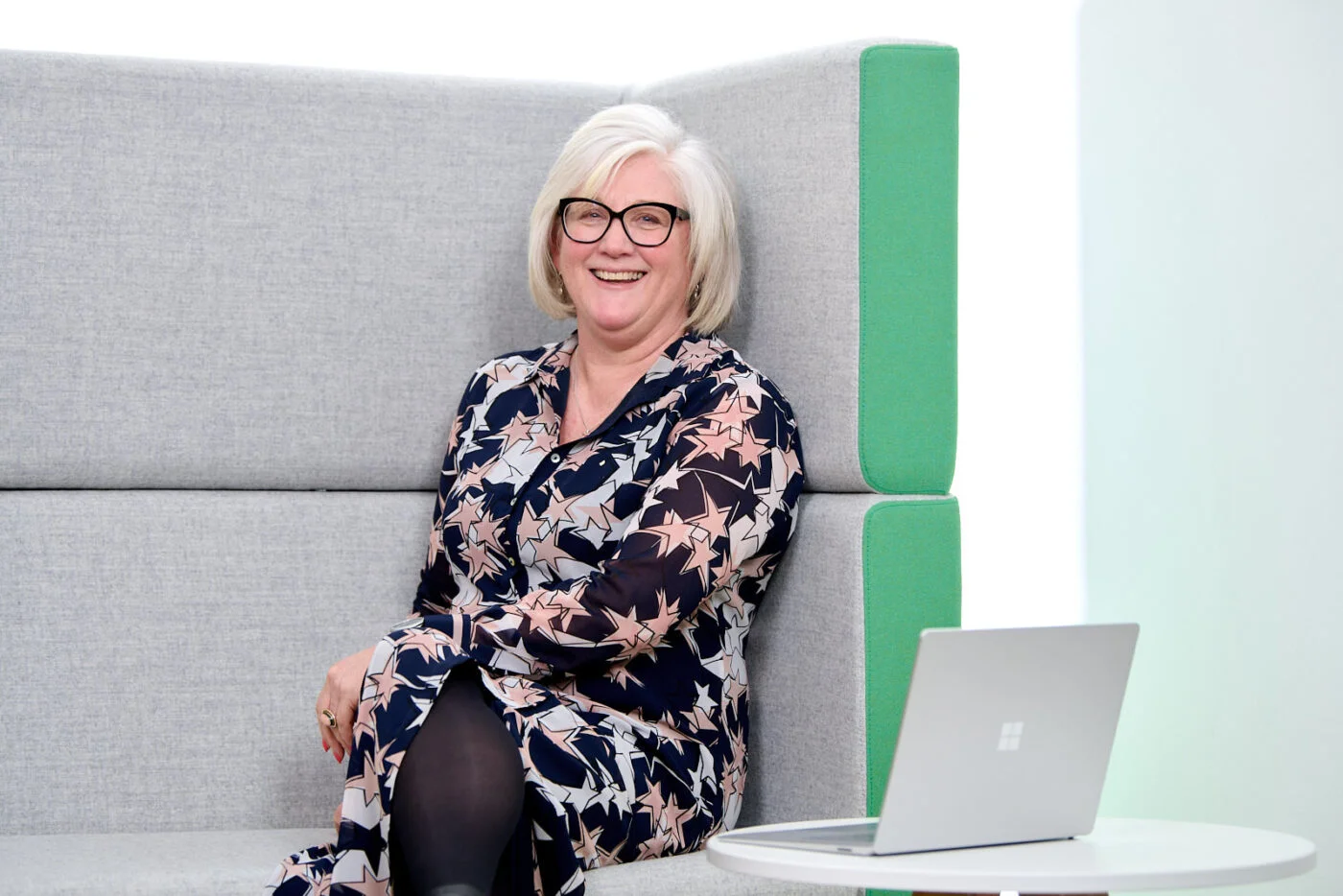 A person with short white hair and glasses, wearing a patterned dress, sits on a gray bench next to a small white table with a laptop. The background is light and has a green cushion on the bench. Spencer Cobby