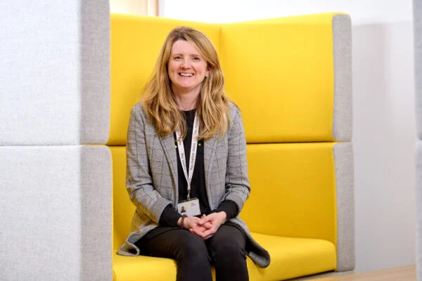 A woman with long hair is seated on a bright yellow cushioned bench. She is wearing a gray blazer and has a lanyard around her neck, smiling at the camera. The surroundings are minimalistic with gray and yellow panels. Spencer Cobby