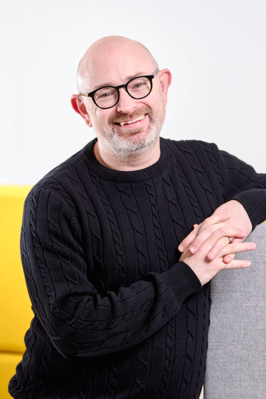 A smiling man with glasses and a beard wearing a black cable-knit sweater is leaning on a gray armrest. The background features a yellow sofa. Spencer Cobby