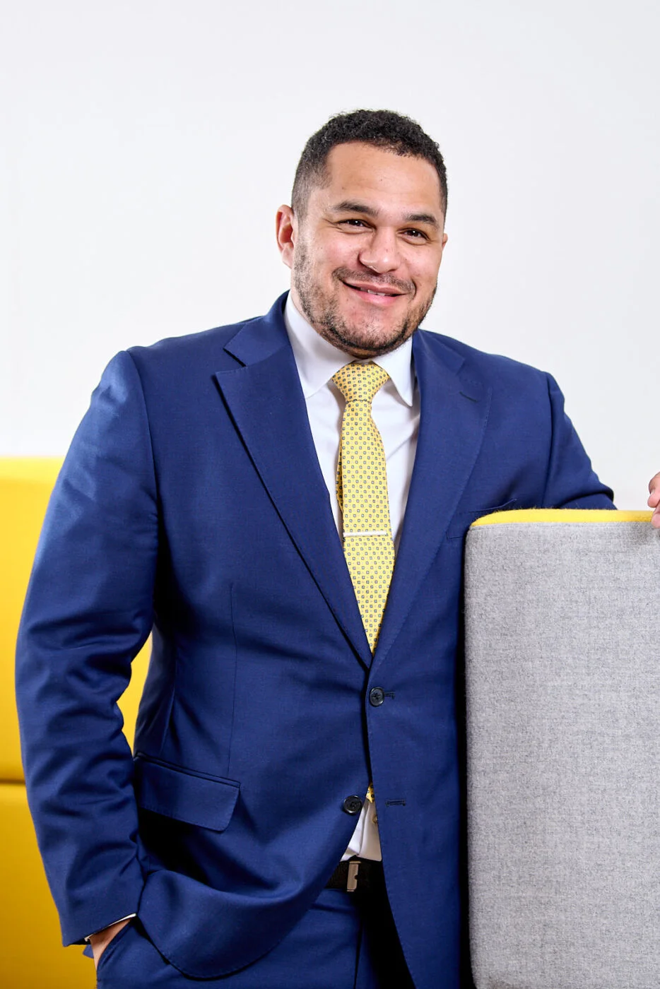 Man in a blue suit with a yellow tie leans against a gray chair, smiling. The background features bright yellow accents. Spencer Cobby